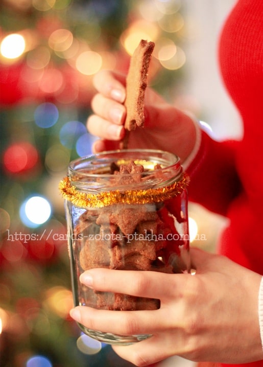 Hand hält einen Lebkuchen-Stern über einem mit goldener Lametta umwickelten Glas, Weihnachtsbaum im Hintergrund