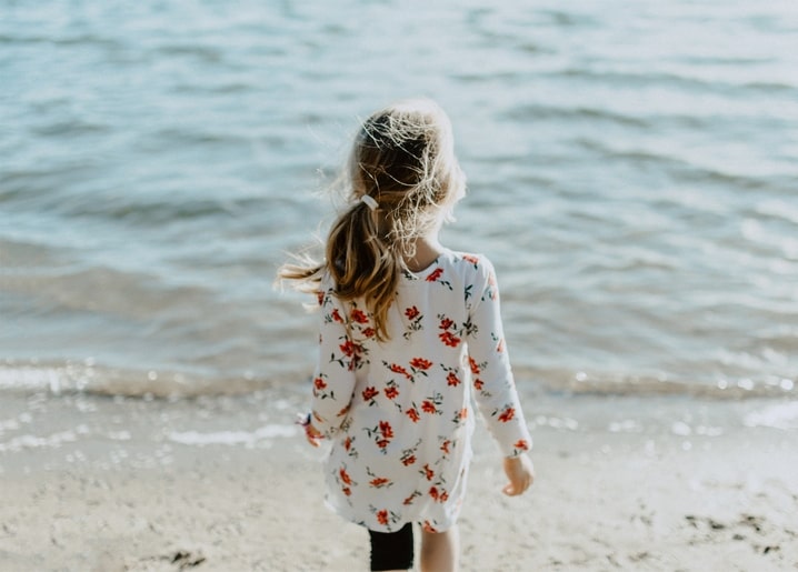 Mädchen im geblümten Kleid geht barfuß am Wasser entlang, Haare im Wind