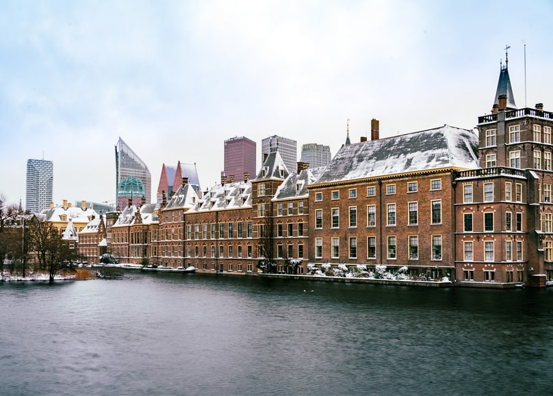 Snow-covered Binnenhof buildings in The Hague beside a canal, modern skyline behind