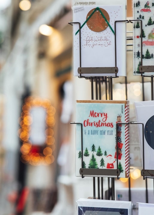 Christmas greeting cards on a display rack in a shop window, lit by warm bokeh string lights.
