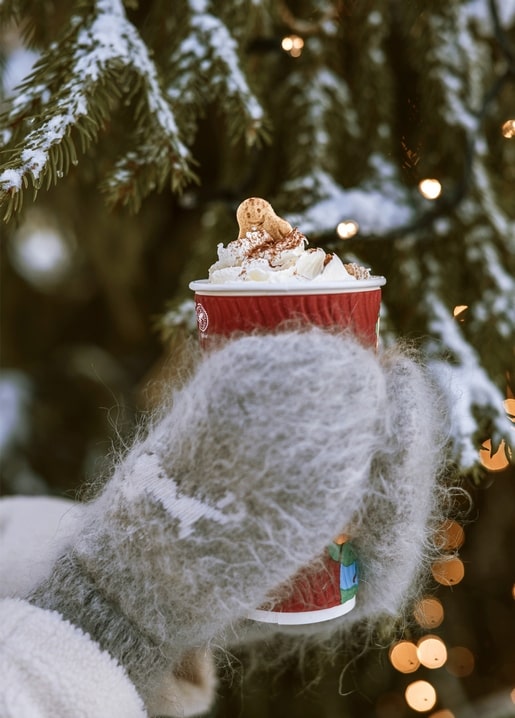Hand im Fäustling hält heiße Schokolade mit Sahne und Lebkuchenmann vor Tannenbaum