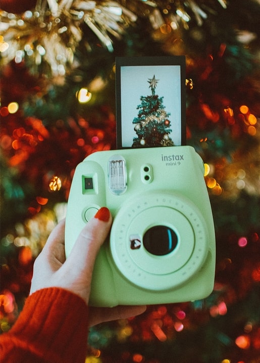 Hand im roten Pullover hält eine Instax-Kamera, die ein Weihnachtsbaum-Foto druckt