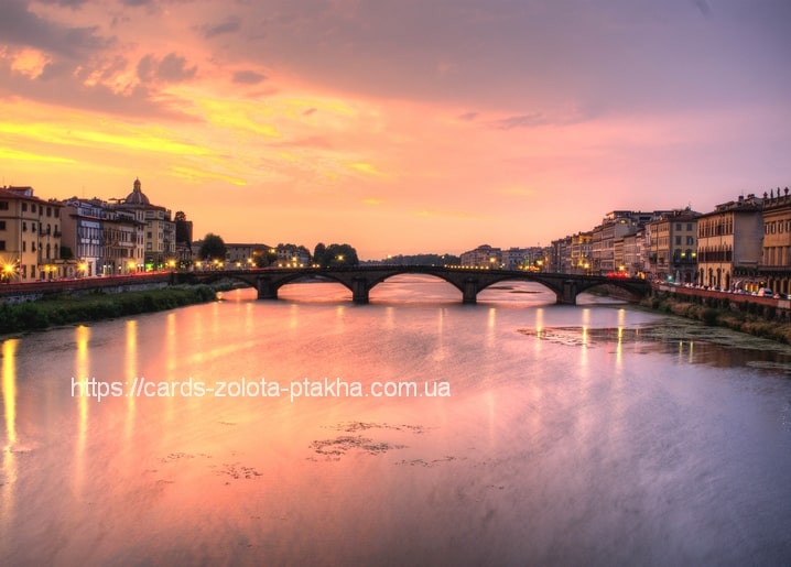Steinbogenbrücke über einem italienischen Fluss bei Sonnenuntergang mit Häuserreflexionen im Wasser.