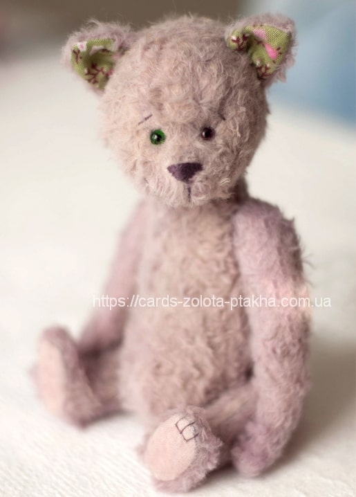 Mauve mohair teddy bear with one green and one brown eye, sitting on white fabric