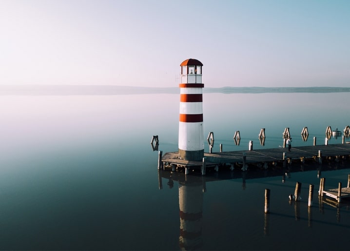 Rot-weißer Leuchtturm spiegelt sich im ruhigen See neben einem Holzsteg bei Sonnenaufgang