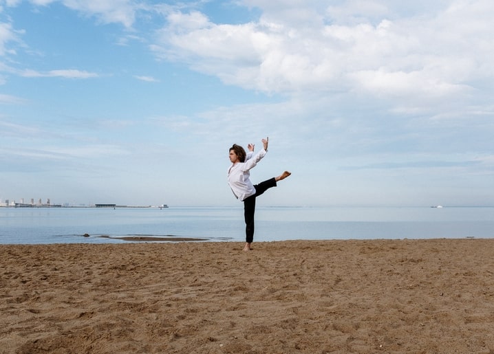 Mann im weißen Hemd vollführt einen hohen Kick am Strand vor einer Stadt-Skyline