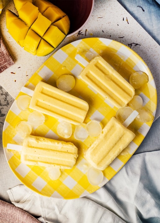 Gelbes Mango-Eis am Stiel auf kariertem Teller mit Mangowürfeln und Eis