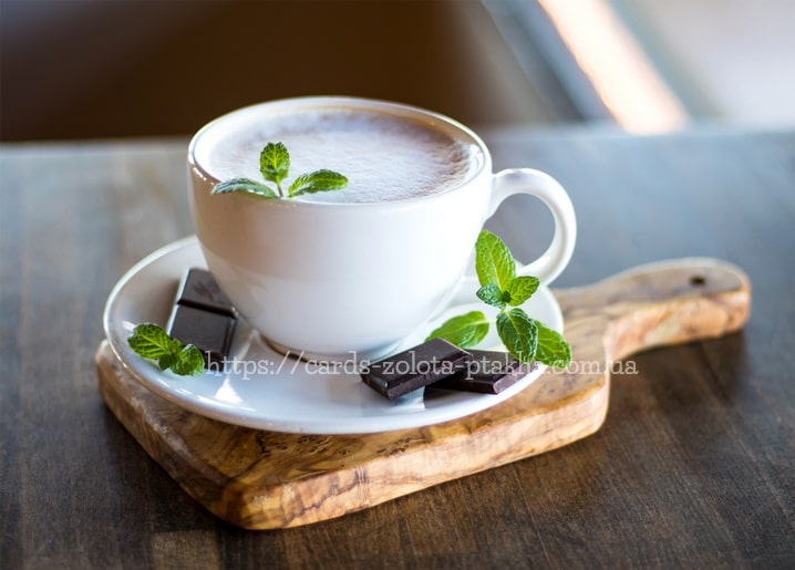 Weißer Cappuccino mit Minzblatt-Deko neben dunklen Schokoladenstückchen auf einem Holzbrett