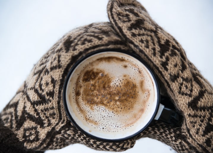 Gestrickte Fäustlinge mit Norwegermuster halten einen Emaillebecher mit Kaffeeschaum im Schnee