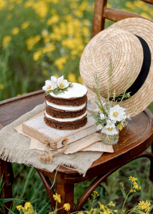 Naked Cake mit weißen Blüten neben Strohhut auf altem Buch in der Wiese