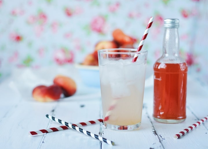 Glas Pfirsich-Eistee mit gestreiften Strohhalmen, Pfirsichen und einer Flasche Limonade vor Blumentapete