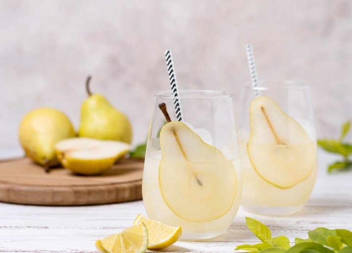 Zwei Gläser Birnenlimonade mit gestreiften Strohhalmen neben frischen Birnen und Zitrone.