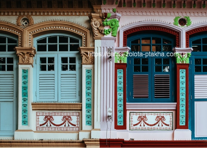 Zwei gleiche Peranakan-Fenster nebeneinander: links zartes Türkis, rechts tiefes Petrolblau mit weinrotem Fliesenrahmen.