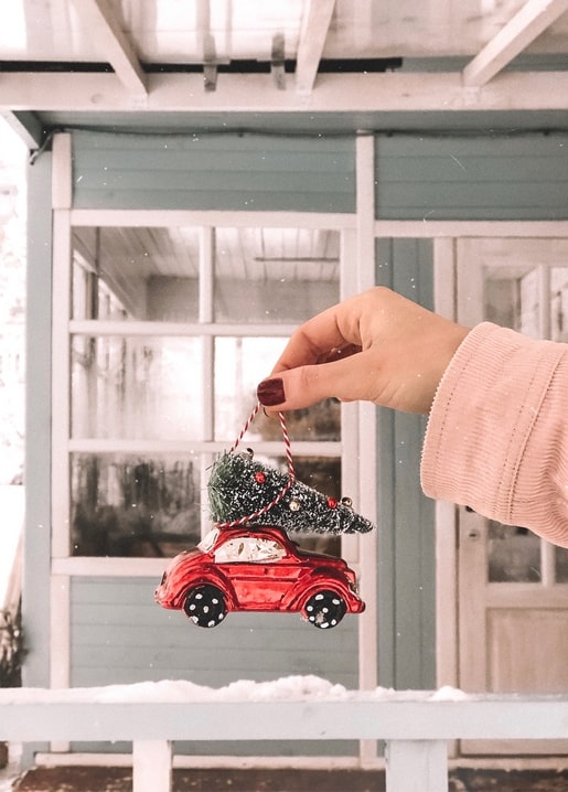 Hand hält ein rotes Glasauto mit Weihnachtsbaum auf dem Dach vor einer Veranda