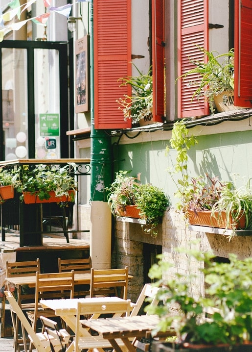 Straßencafé mit roten Fensterläden, Blumentöpfen und leeren Holzstühlen