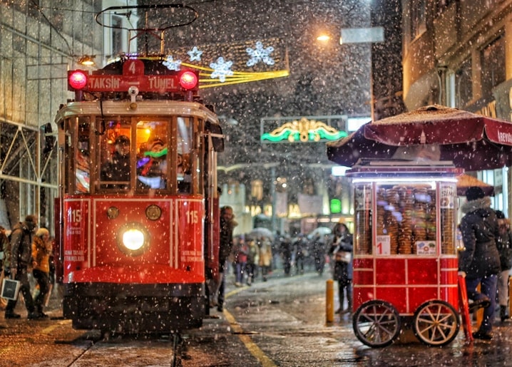 Vintage red tram glowing at night in falling snow beside a lit street-food cart