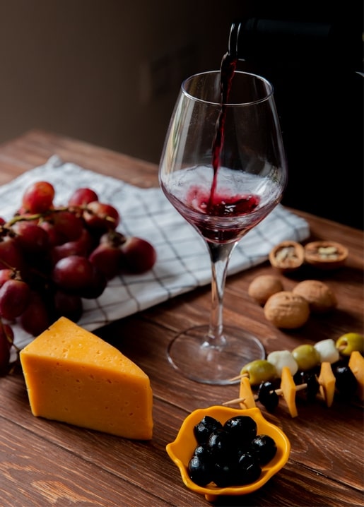 Red wine pouring into a glass beside cheese, grapes, walnuts and olive skewers