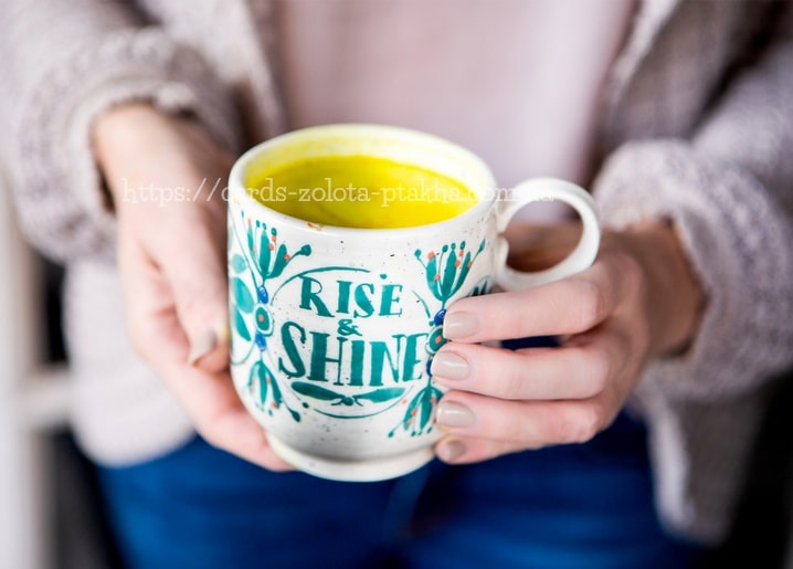 Hände halten eine türkise Volkskunst-Tasse mit Aufschrift „Rise & Shine“ und goldenem Getränk