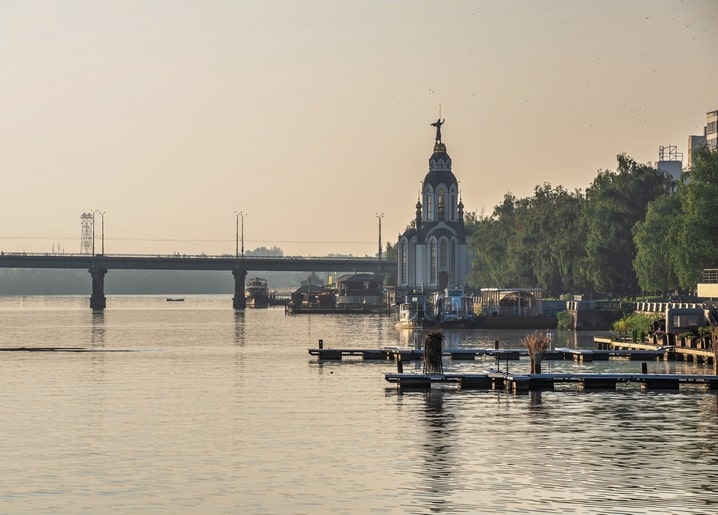 Невелика каплиця на березі Дніпра з човнами і мостом на світанку
