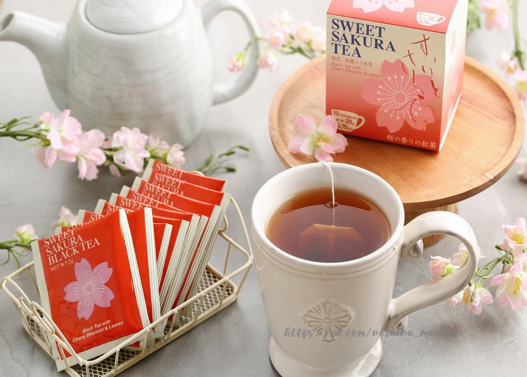 Box mit Sakura-Schwarztee neben weißer Teekanne, Teebeuteln und Kirschblütenzweigen