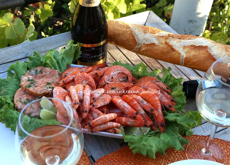 Grilled shrimp with lettuce and lime beside a baguette and two glasses of wine on an outdoor wooden table