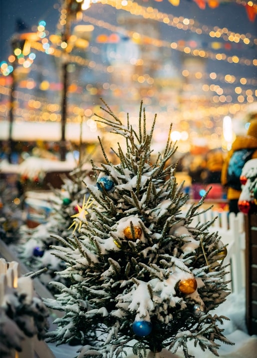Snow-covered Christmas tree decorated with baubles and string lights at an outdoor winter market