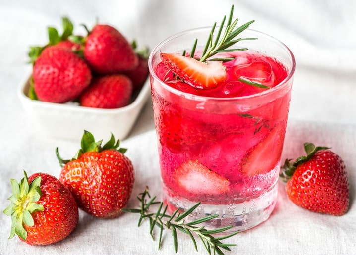 Glas rosa Erdbeer-Limonade mit Eis und Rosmarin, umgeben von frischen Erdbeeren