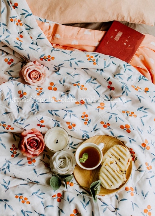 Tee und getoastetes Sandwich zwischen Stoffrosen und Kerzen auf floral gemusterter Bettwäsche