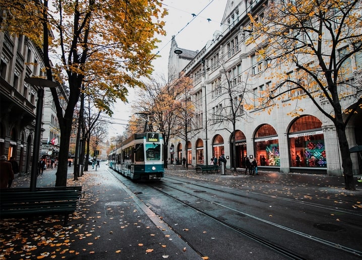 A tram passing along a European city street lined with autumn trees and shopfronts