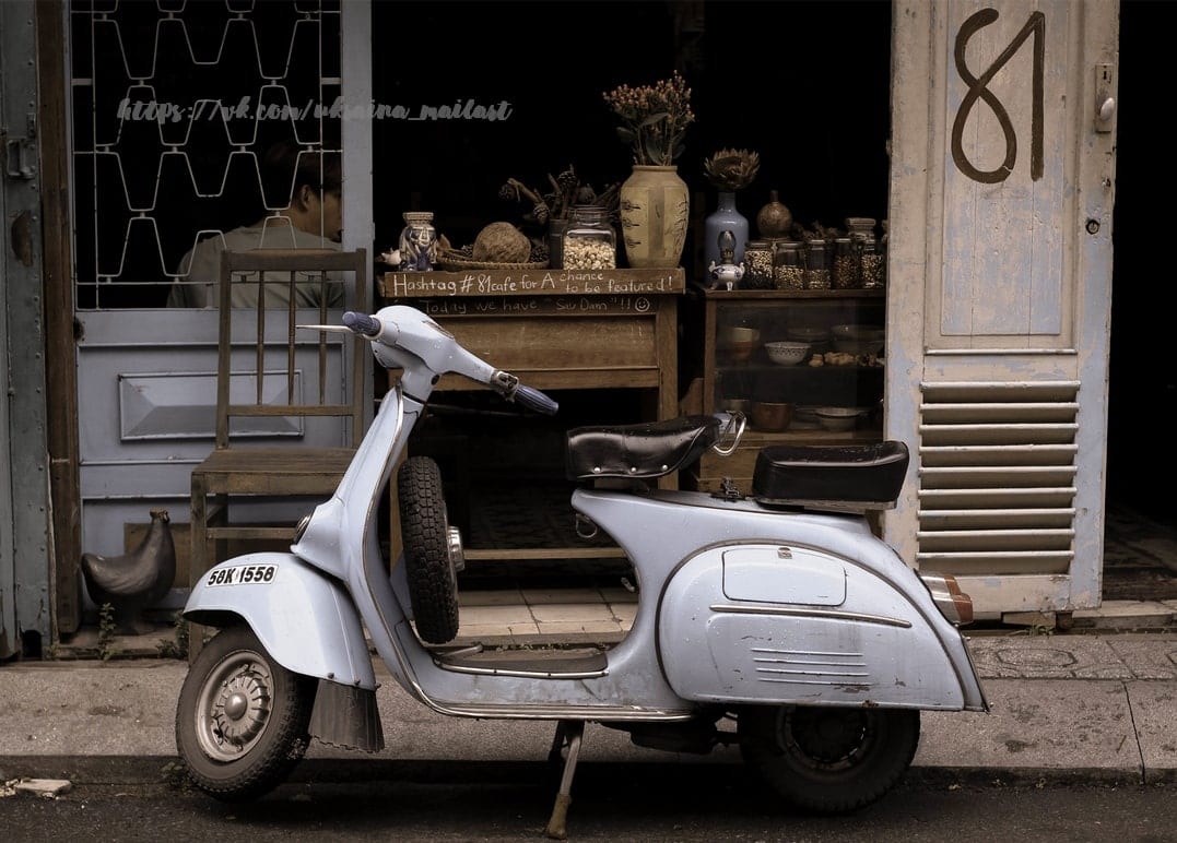 Pale blue vintage Vespa scooter parked outside a curiosity shop numbered 81
