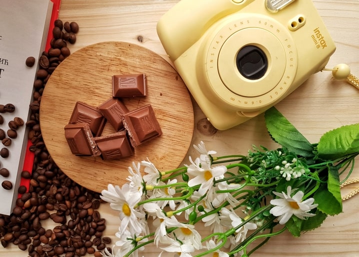 Gelbe Sofortbildkamera, Schokoladenstücke, Kaffeebohnen und Kamillenblüten auf einem Holztisch