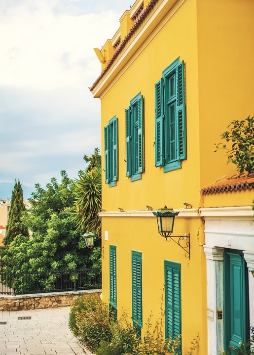 Sonnenbeschienenes gelbes Haus mit türkisen Fensterläden und Laterne an einer Zypressengasse