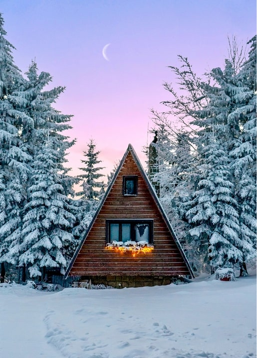 Snow-covered A-frame cabin glowing with lights under a crescent moon at twilight