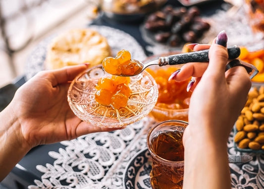 Hand taucht einen Löffel in goldene Marmelade über einer Kristallschale, daneben Tee, Datteln und Mandeln