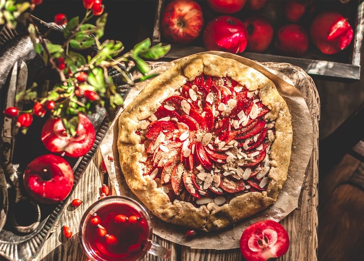 Apfelgalette mit Mandeln, Hagebutten, Tee und einer Kiste roter Äpfel auf einem Holztisch