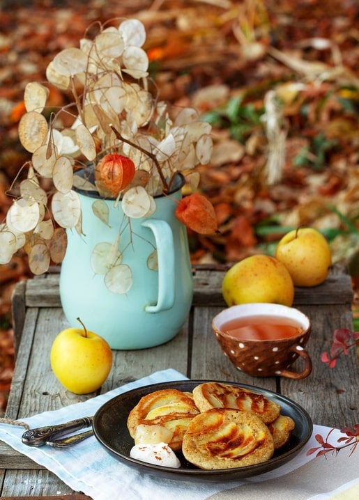 Türkiser Krug mit Trockenblumen, gelbe Äpfel, Tee und ein Teller Apfelpfannkuchen