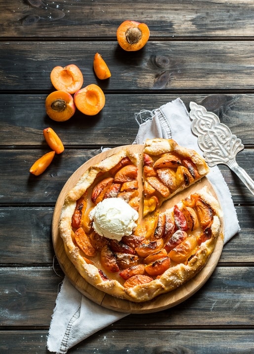 Apricot galette topped with vanilla ice cream beside halved fresh apricots on a wooden table