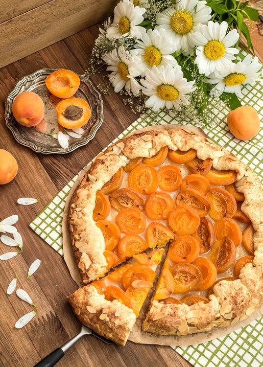 Apricot tart topped with sliced almonds beside a daisy bouquet on a wooden table