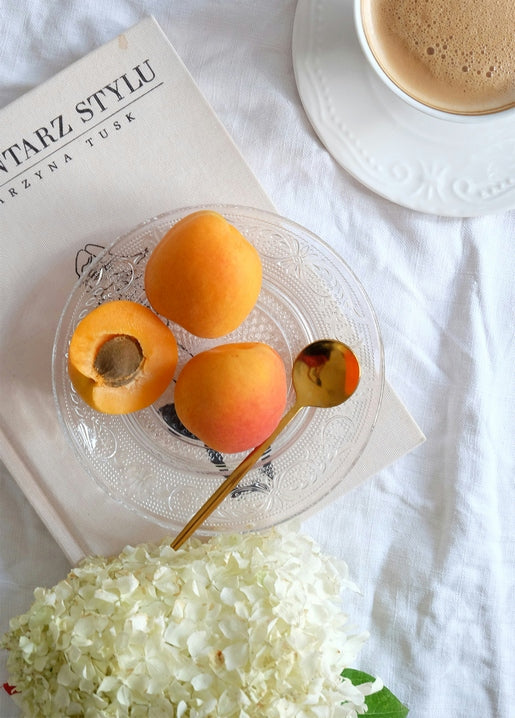 Flat-lay of halved apricots, coffee, white hydrangea and a book on white linen, styled for a summer postcard.
