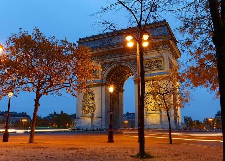 Arc de Triomphe illuminated at night in Paris, framed by autumn trees and traffic light trails