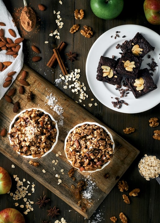 Draufsicht auf Mandel-Brownies, Apfel-Nuss-Crumble, Walnüsse und Zimt auf Holz
