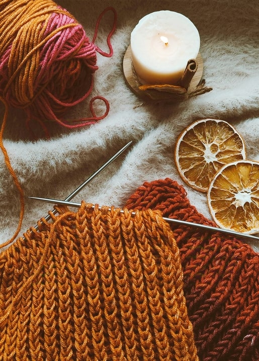 Orangefarbene und rosa Wolle, Stricknadeln, brennende Kerze und getrocknete Orangenscheiben auf einer Strickdecke