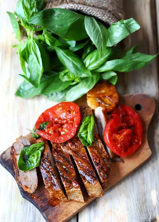 Food postcard featuring sliced grilled pork with ripe tomatoes and fresh basil on a wooden board