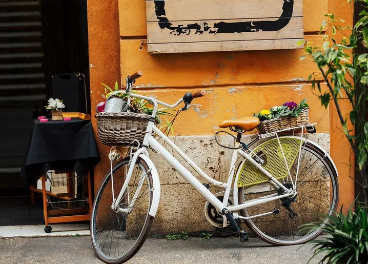 European street photo postcard featuring a white bicycle with wicker flower baskets beside a terracotta wall