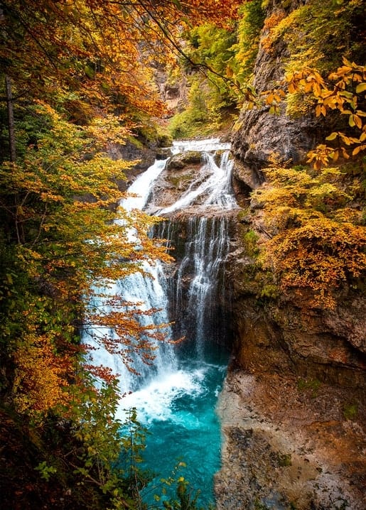 Wasserfall stürzt in ein türkisfarbenes Becken in herbstlicher Waldschlucht