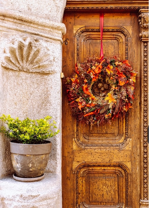 Herbstkranz aus trockenem Laub und Zapfen an einer geschnitzten Holztür neben einem Blumentopf