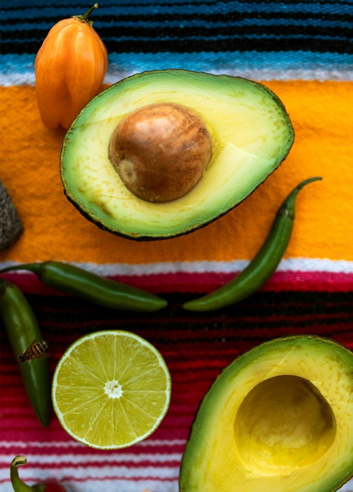 Halved avocado, cut lime and green and orange chili peppers arranged on a colorful woven blanket.
