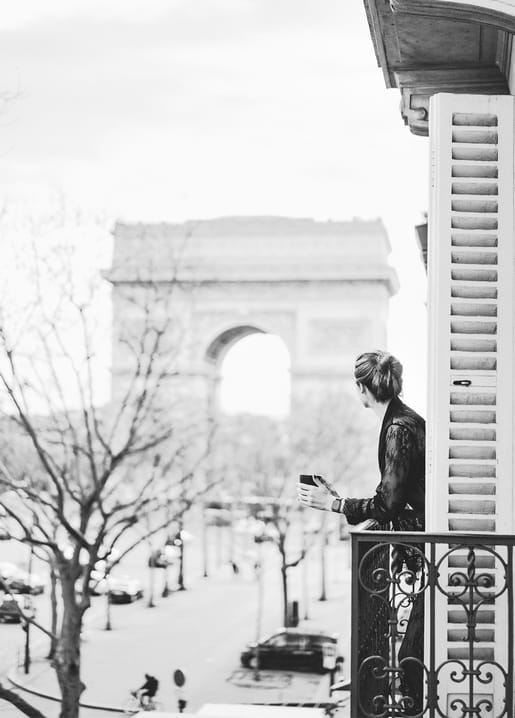 A woman on a wrought-iron balcony holds a coffee cup while looking toward the Arc de Triomphe.