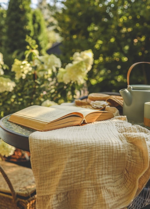 Aufgeschlagenes Buch und Teekanne auf leinenbedecktem Gartentisch im Sonnenlicht