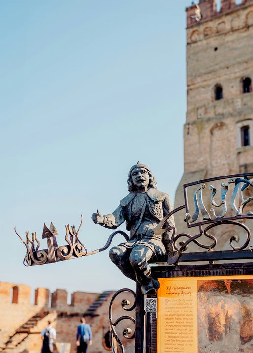 Bronzestatue in historischer Tracht auf einem kunstvollen Lutsk-Schild, Burgturm unscharf im Hintergrund
